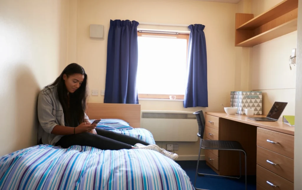 Person-sitting-on-bed-in-supported-accommodation-room-with-blue-striped-bedding-and-modern-furniture